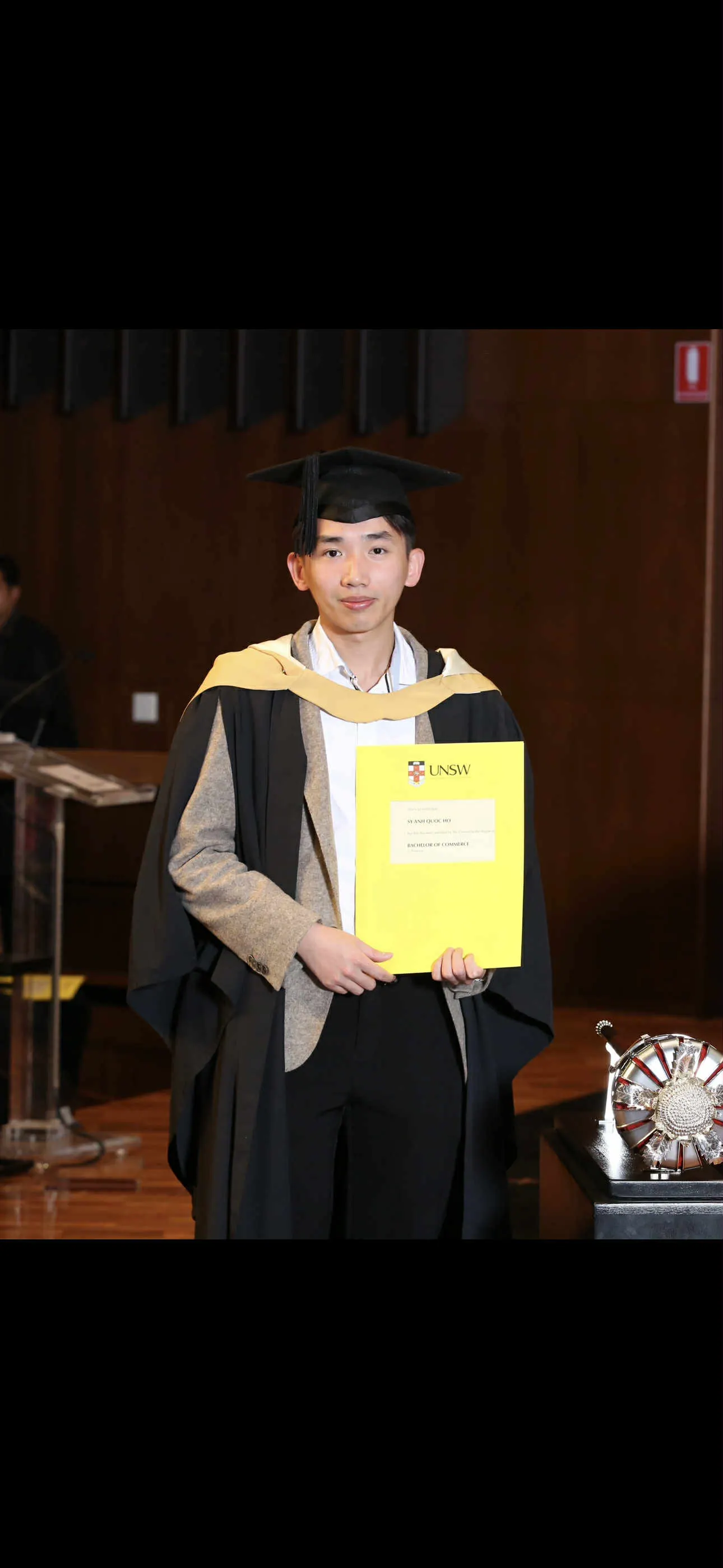 Johnny in graduation gown and cap holding his UNSW testamur
